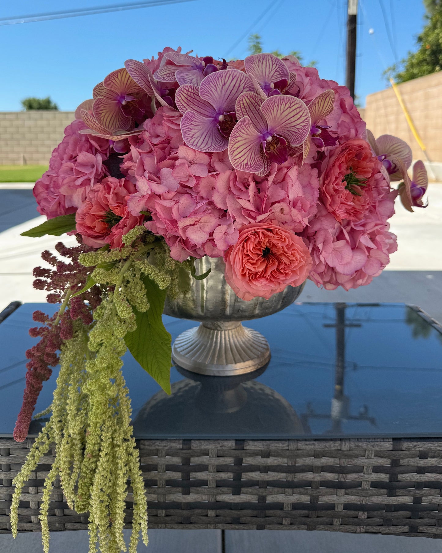 Bouquet of pink flowers in a silver vase on a reflective surface with a blurred outdoor background.