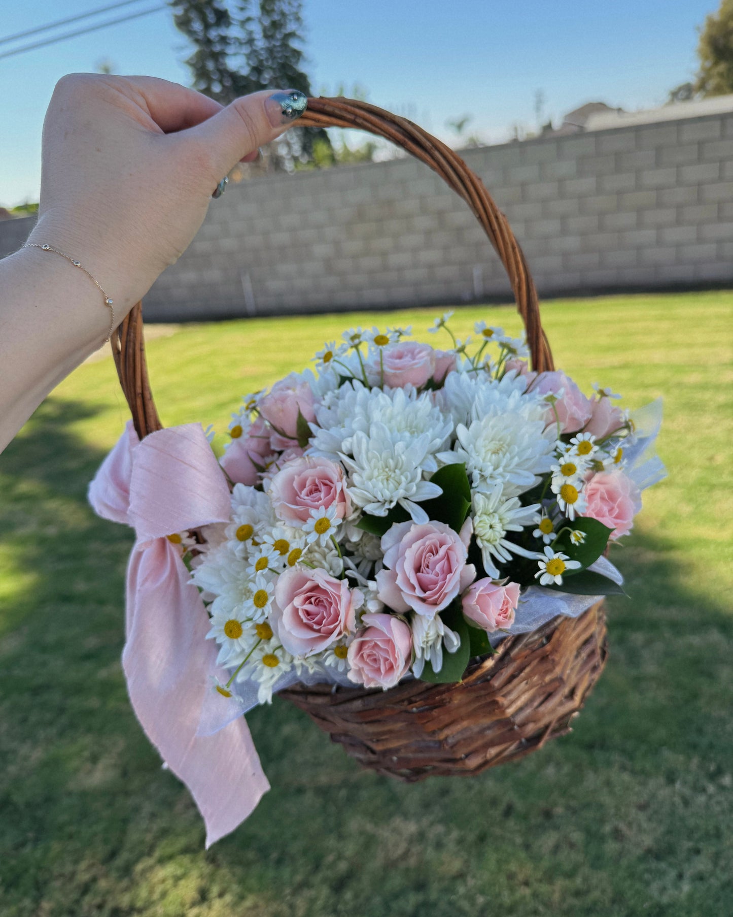 Hand holding a basket of flowers with a pink ribbon against a grassy background