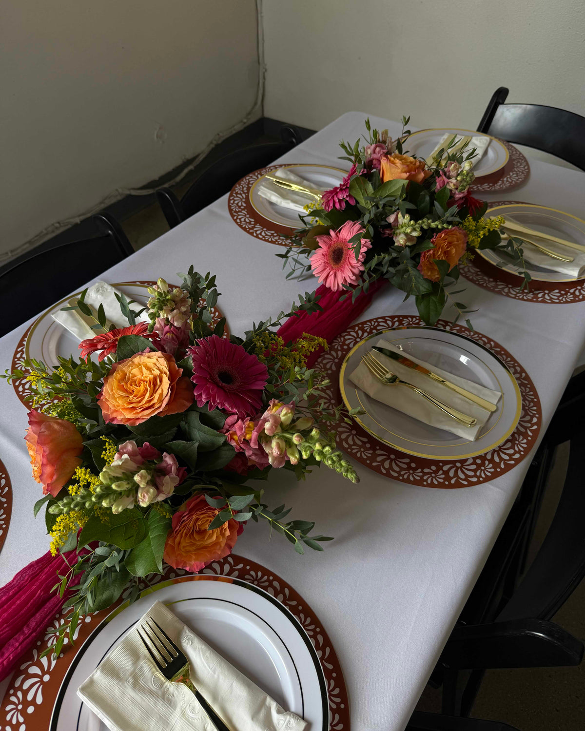 A styled dining table set with multiple floral centerpieces, white tablecloths, patterned brown and gold plates, folded cream napkins, and gold-toned cutlery, photographed in a real indoor environment with chairs visible.