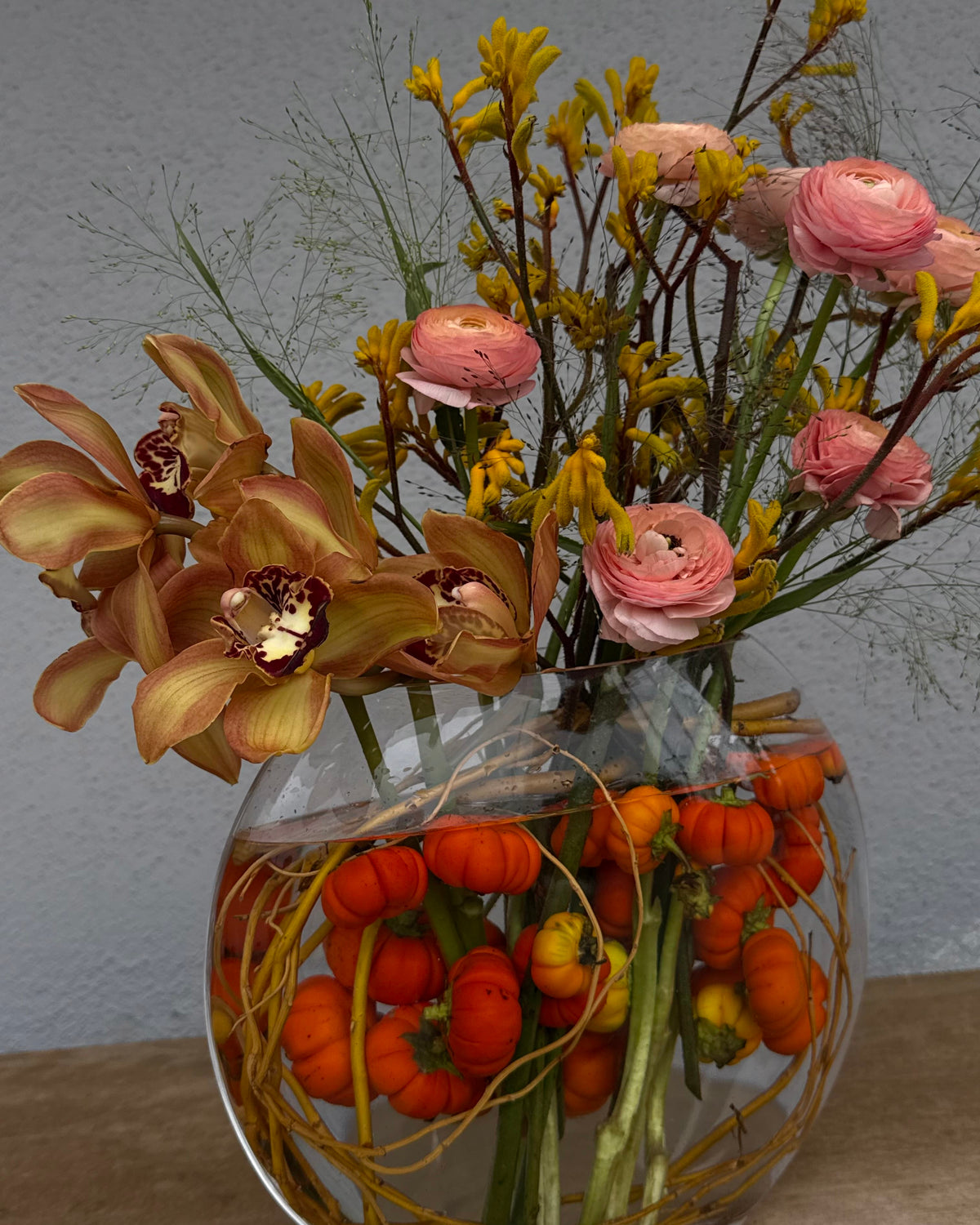 An autumnal floral arrangement in a round glass fishbowl vase, featuring pink ranunculus, yellow wildflowers, and tan orchids with orange mini pumpkins floating in the water, set against a neutral gray wall.