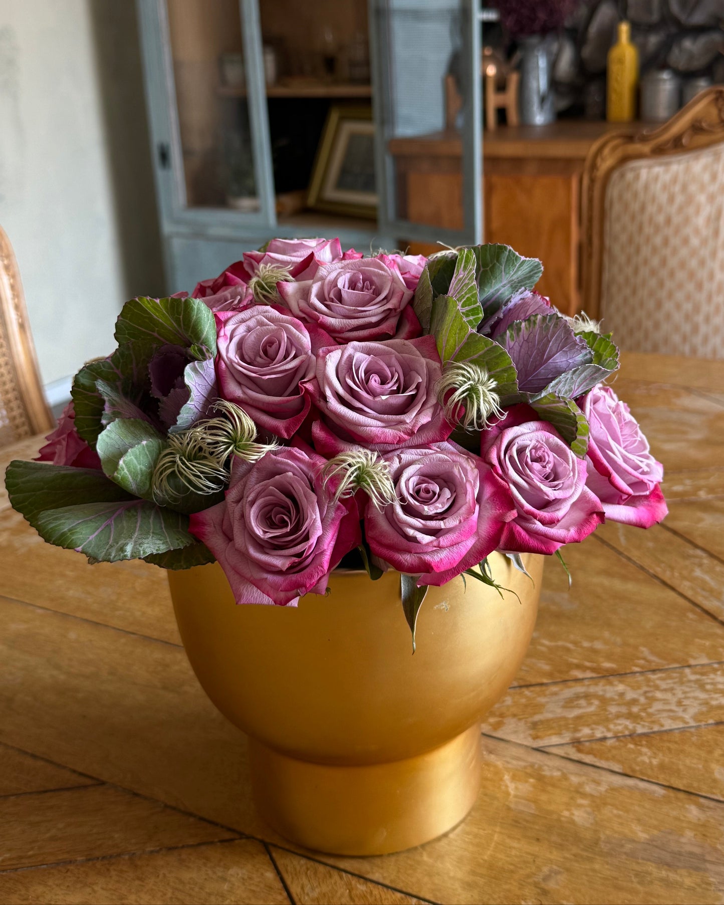 lavender, decorative cabbage, and soft filler thoughtfully arranged in a gold vase and placed on a wooden table.