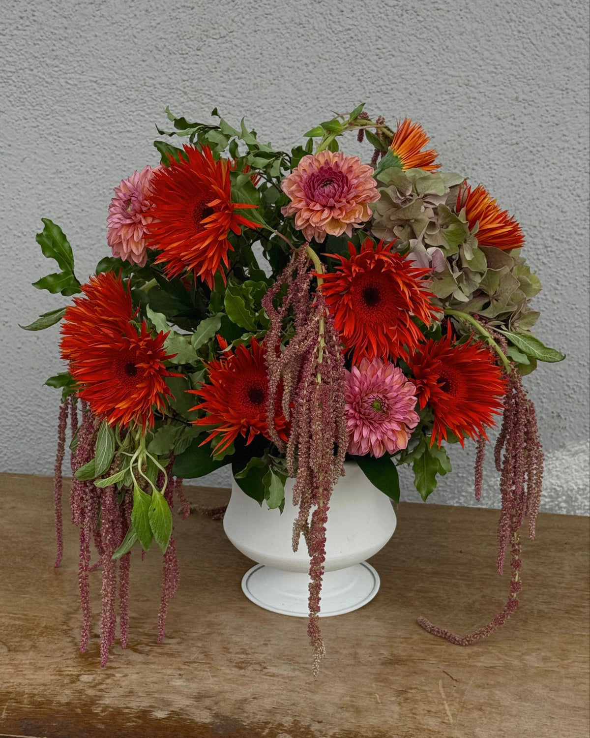 A vibrant autumnal flower arrangement featuring red-orange gerbera daisies, orange dahlias, green antique hydrangea ans foliage, and hanging burgundy amaranthus, presented in a white ceramic pot on a rustic wooden surface against a textured gray wall.