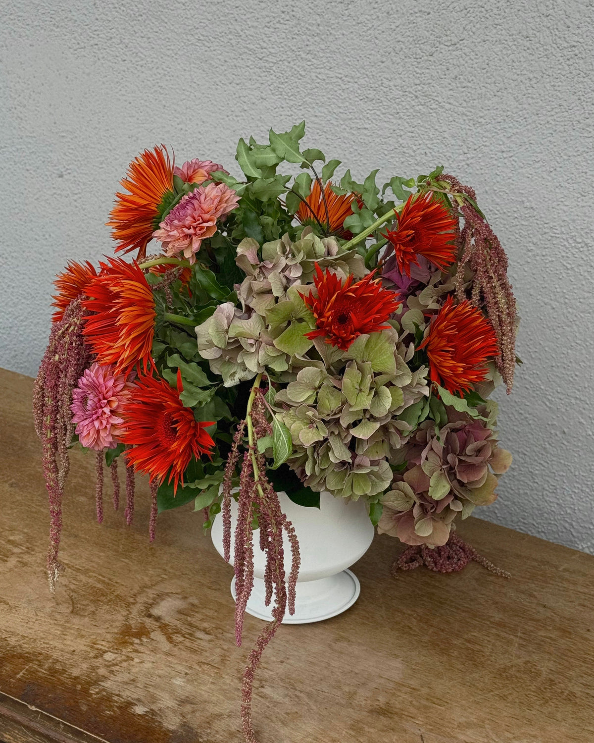 A vibrant flower arrangement in a white pedestal vase, featuring bright orange-red gerbera daisies, dahlias, and green hydrangeas with trailing burgundy stems, displayed on a rustic wooden table against a textured gray wall.