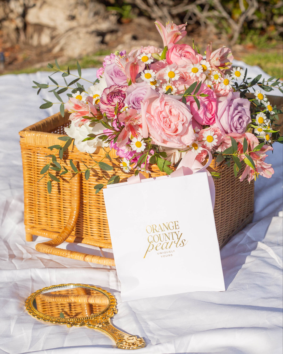 A pink and white floral arrangement in a wicker basket on a white draped blanket, with a white card reading 'Orange County Pearls' and a small gold-handled mirror in the foreground. The background shows an outdoor garden setting.