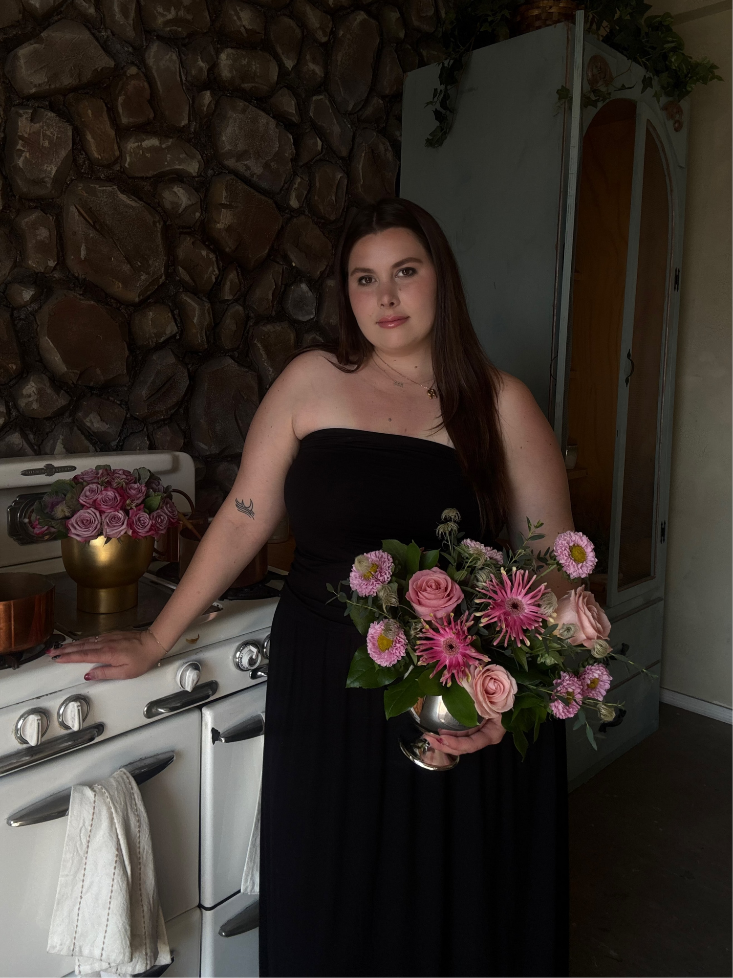 Woman in a black dress holding flowers in a kitchen with stone walls.
