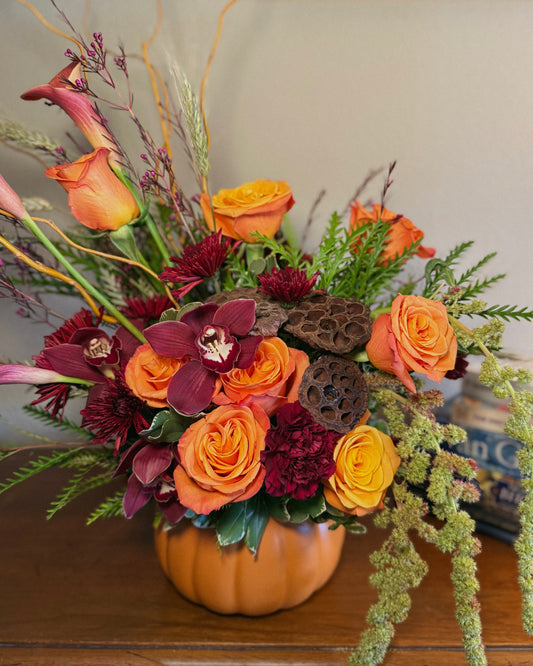 Luxury floral arrangement in a pumpkin vase with orange and red flowers on a wooden surface. Crafted by Superbloom floral. 