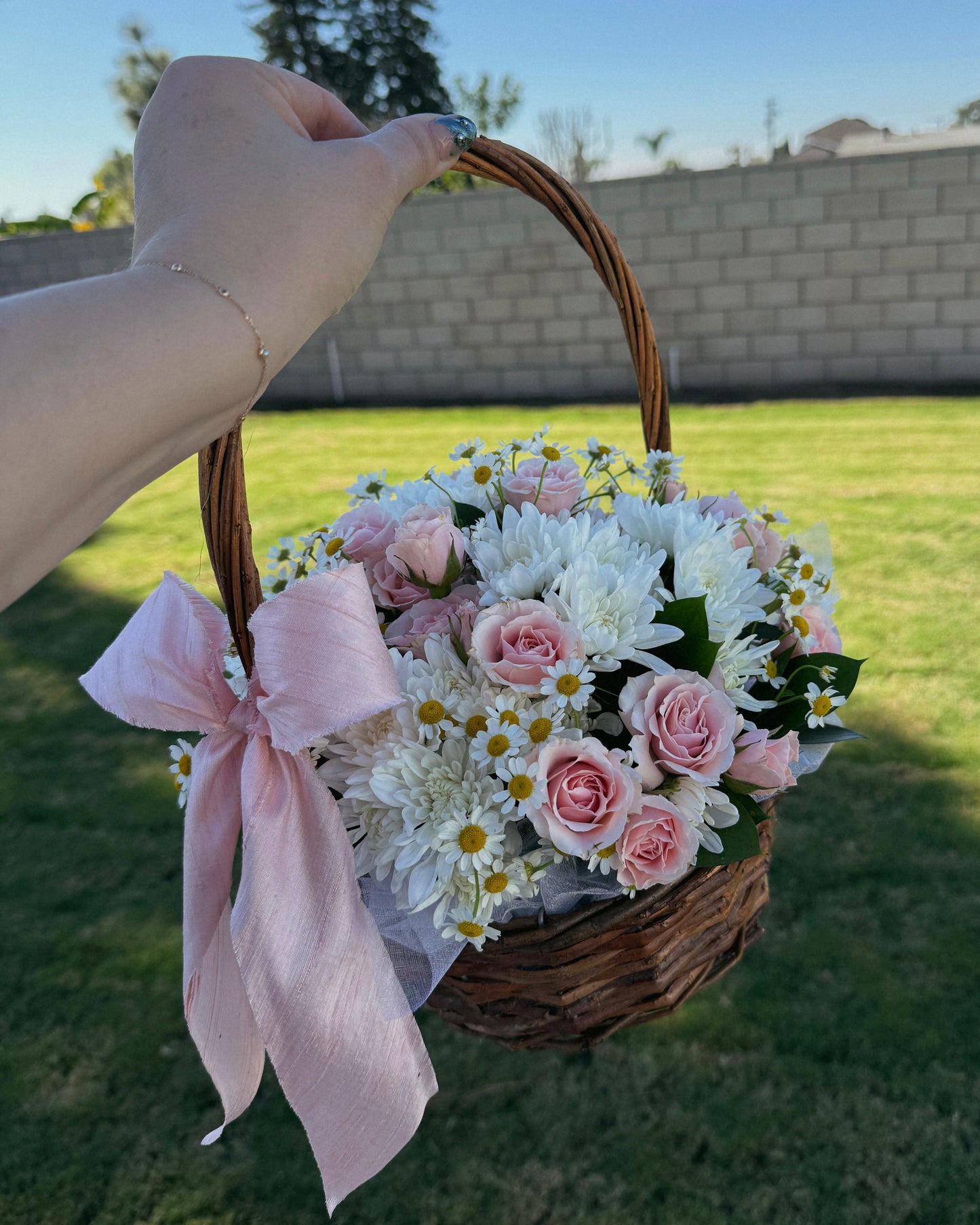 Basket of flowers with a pink ribbon held by a hand outdoors.