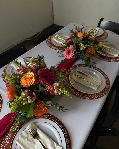 A styled dining table set with multiple floral centerpieces, white tablecloths, patterned brown and gold plates, folded cream napkins, and gold-toned cutlery, photographed in a real indoor environment with chairs visible.