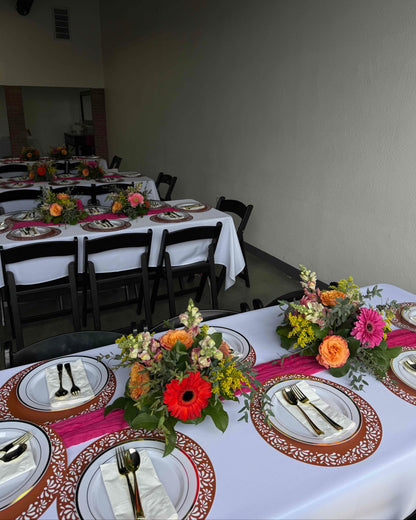 A decorated event table setting with white tablecloths, ornate brown and gold placemats, white plates, folded napkins with gold cutlery, and colorful floral centerpieces with pink, orange, red, and yellow flowers.