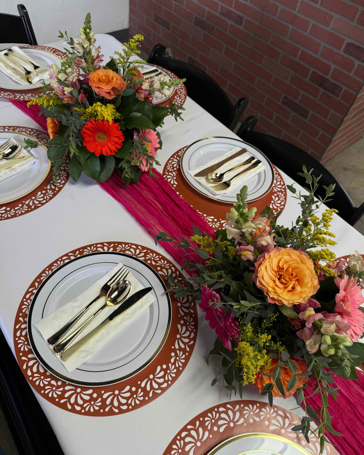 A styled dining table setting with white plates, gold cutlery, orange and pink floral centerpieces, and a bright pink runner against a brick wall background.