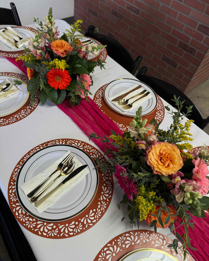 A styled dining table setting with white plates, gold cutlery, orange and pink floral centerpieces, and a bright pink runner against a brick wall background.