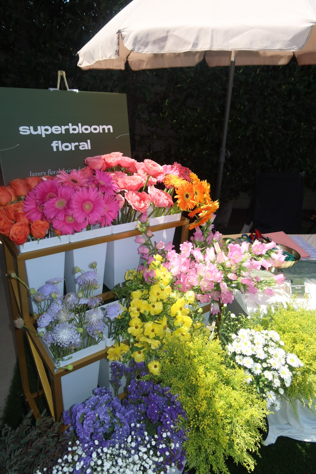 A display of colorful flowers in a cart under a white patio umbrella, with a sign reading 'superbloom floral' on the left. The flowers include pink, orange, yellow, purple, and white blooms in wooden crates. Flower bar experience from superbloom floral.