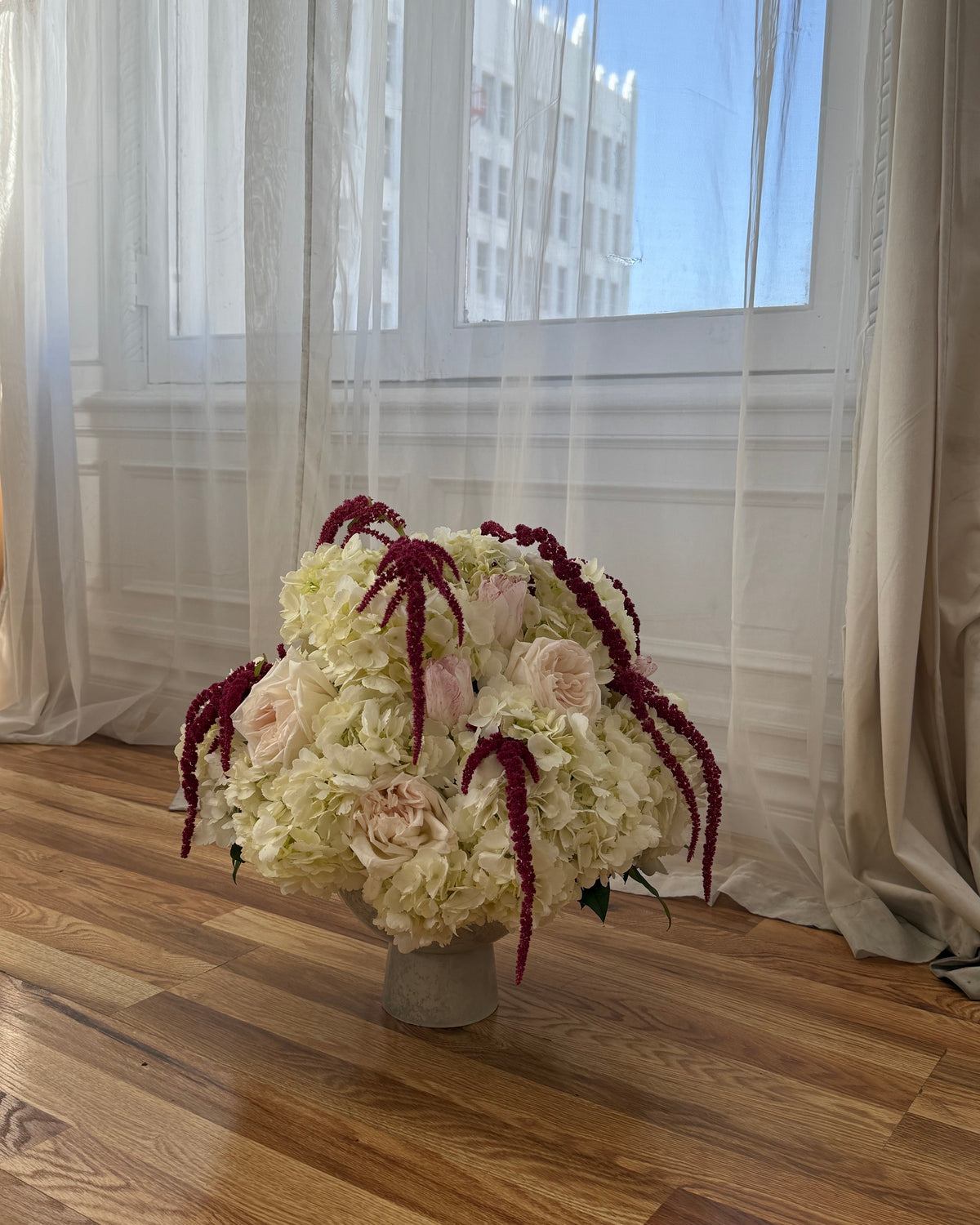 Flower arrangement featuring a lush composition of premium roses, hydrangeas, and delicate seasonal blooms in soft ivory and blush tones, artfully arranged with cascading burgundy textures in a timeless vase. 