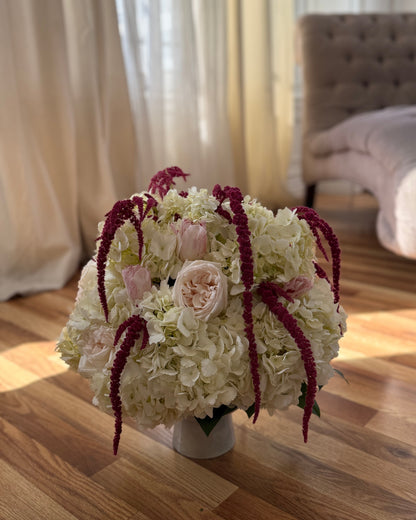 Flower arrangement featuring a lush composition of premium roses, hydrangeas, and delicate seasonal blooms in soft ivory and blush tones, artfully arranged with cascading burgundy textures in a timeless vase. 