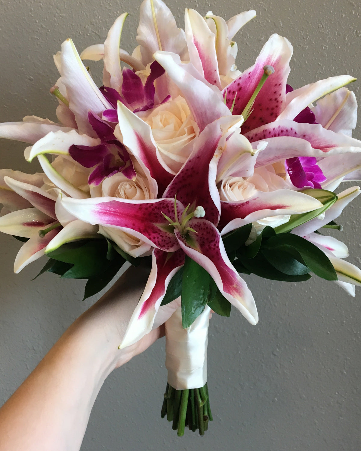 A hand holding a bridal bouquet featuring cream and magenta lilies, white roses, and purple orchids against a neutral gray wall.