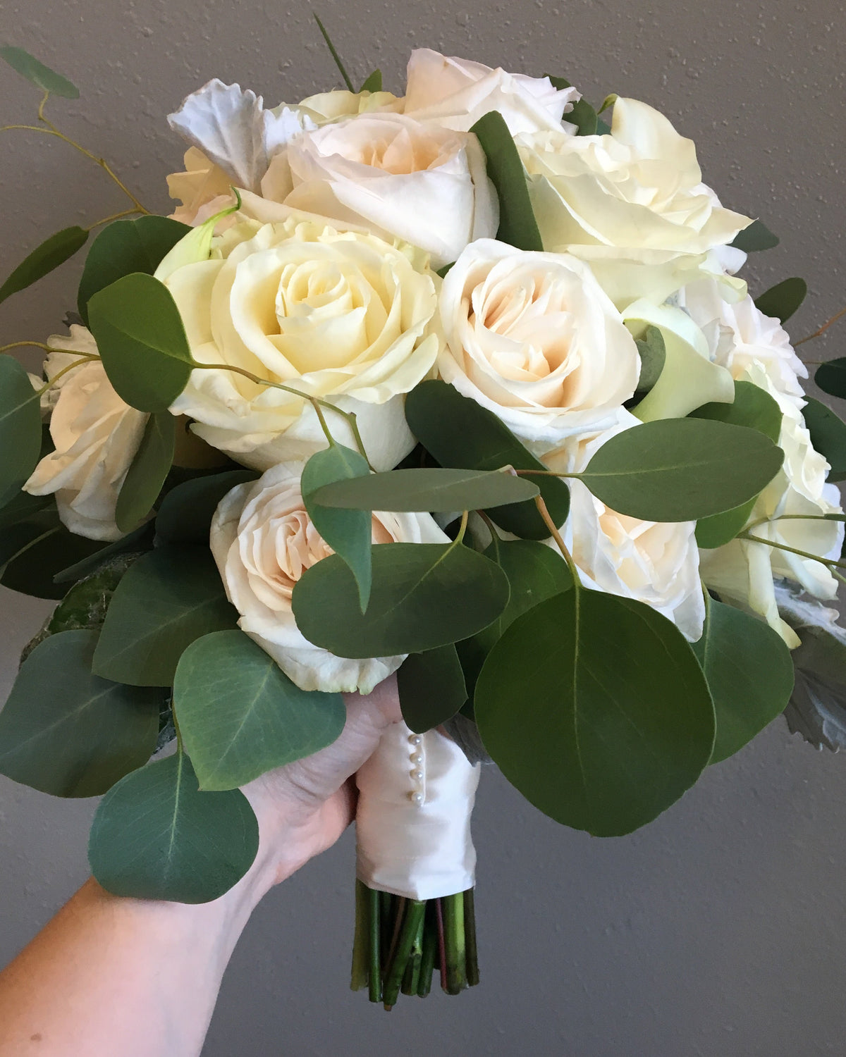 A bridal bouquet featuring cream and off-white roses with green eucalyptus leaves, held by a hand against a neutral gray wall.