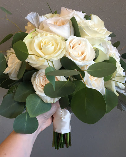 A bridal bouquet featuring cream and off-white roses with green eucalyptus leaves, held by a hand against a neutral gray wall.