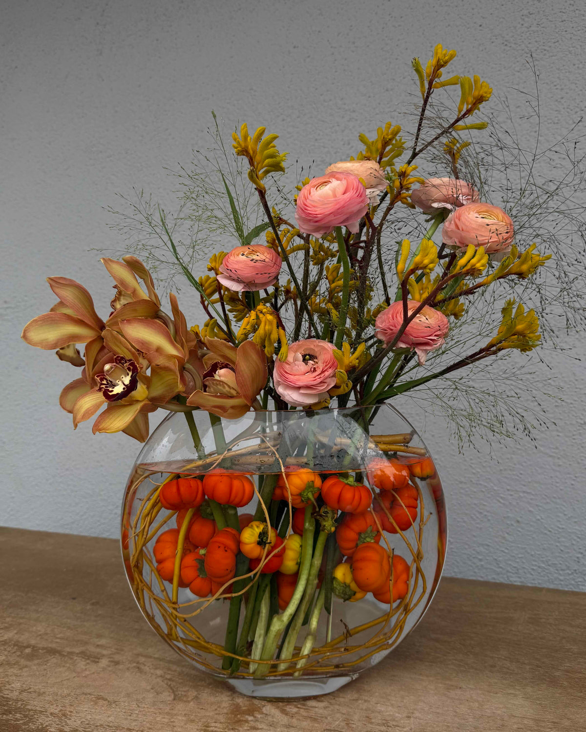 An autumnal floral arrangement in a round glass fishbowl vase, featuring pink ranunculus, yellow wildflowers, and tan orchids with orange mini pumpkins floating in the water, set against a neutral gray wall.
