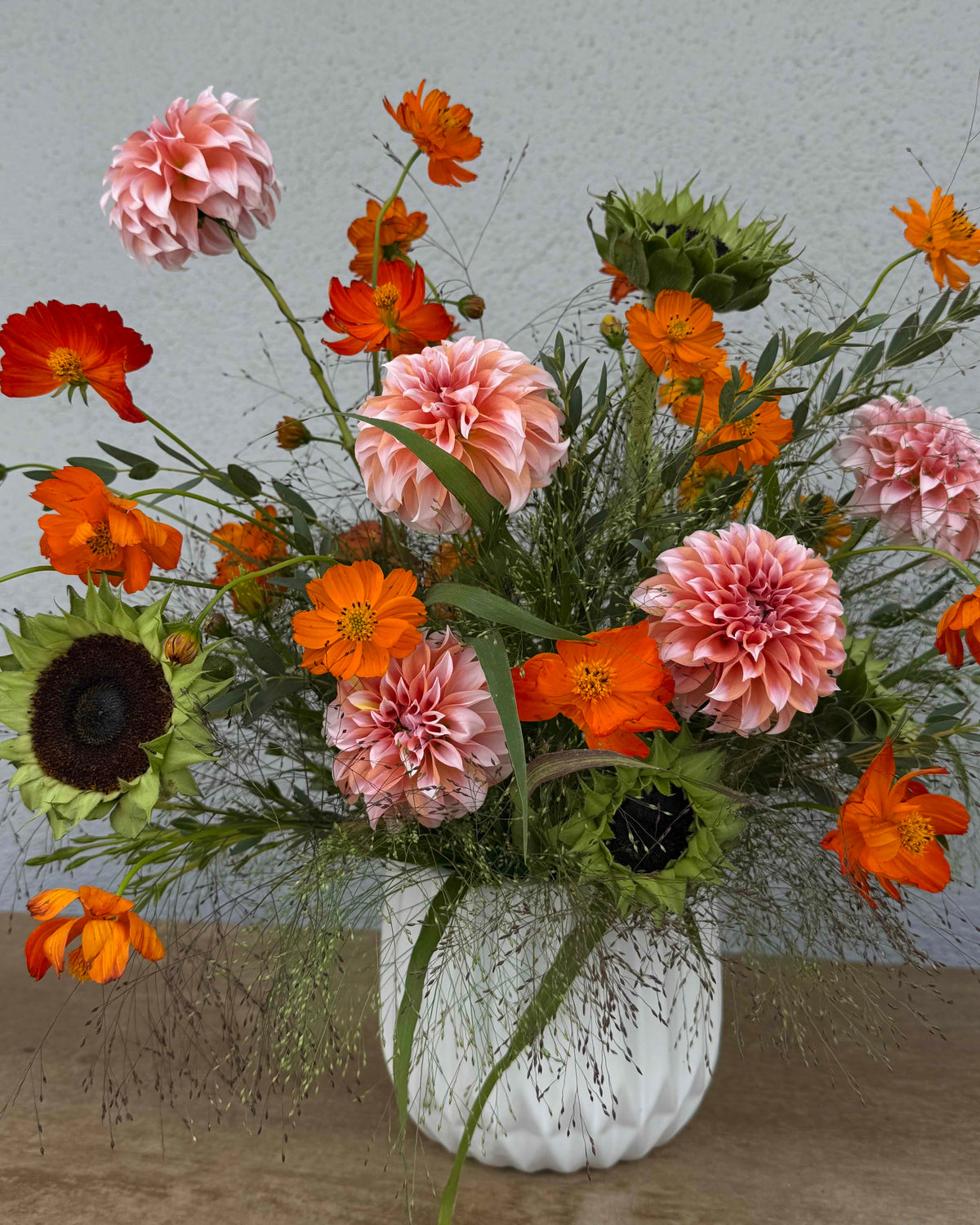 A vibrant floral arrangement featuring pink and orange flowers, including dahlias, cosmos, and sunflowers, styled in a textured white ceramic vase on a rustic wooden surface against a neutral gray wall.