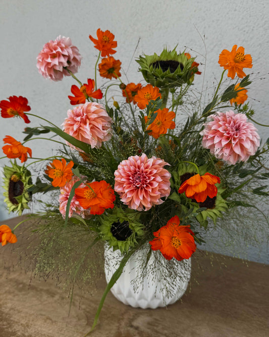A vibrant floral arrangement featuring pink and orange flowers, including dahlias, cosmos, and sunflowers, styled in a textured white ceramic vase on a rustic wooden surface against a neutral gray wall.