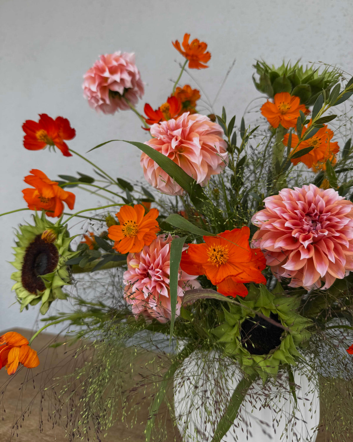 A vibrant floral arrangement featuring pink and orange flowers, including dahlias, cosmos, and sunflowers, styled in a textured white ceramic vase on a rustic wooden surface against a neutral gray wall.