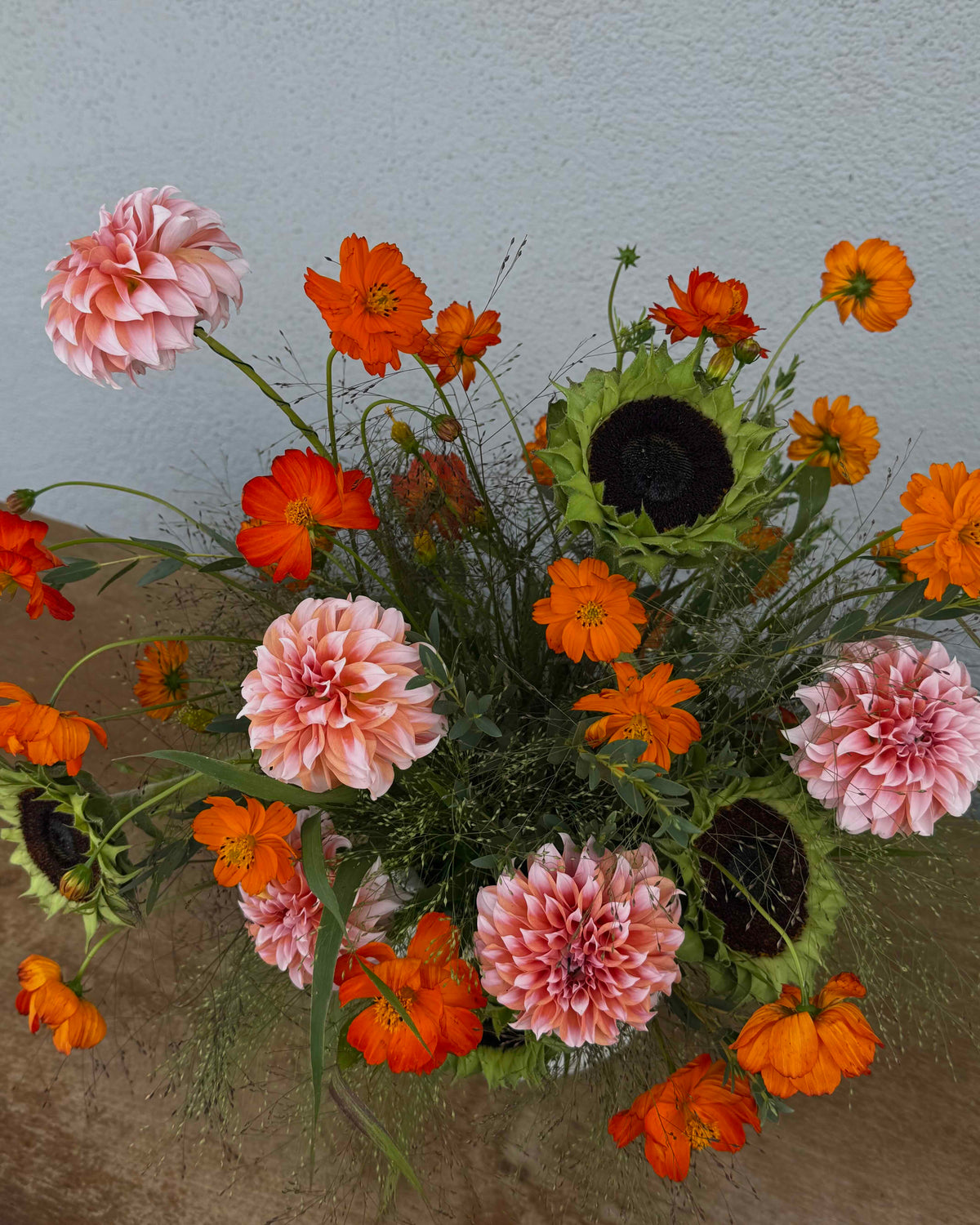 A vibrant floral arrangement featuring pink and orange flowers, including dahlias, cosmos, and sunflowers, styled in a textured white ceramic vase on a rustic wooden surface against a neutral gray wall.