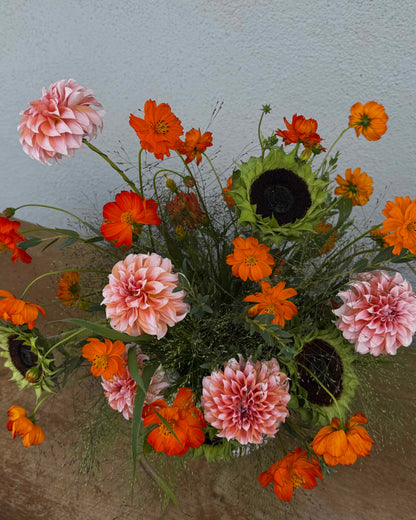 A vibrant floral arrangement featuring pink and orange flowers, including dahlias, cosmos, and sunflowers, styled in a textured white ceramic vase on a rustic wooden surface against a neutral gray wall.