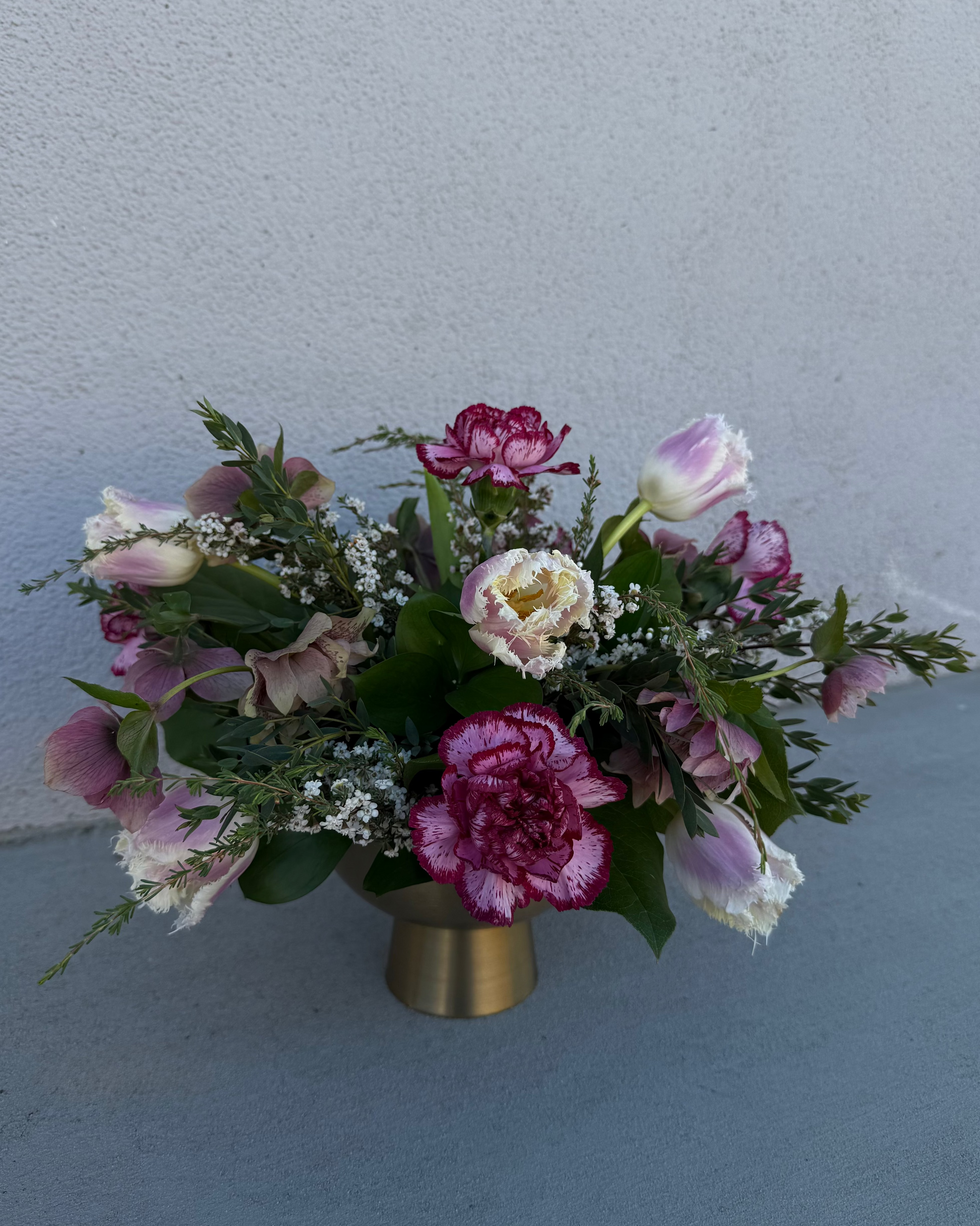 Floral arrangement made from tulips, carnations, and helebore with greenery and filler in a gold vase.
