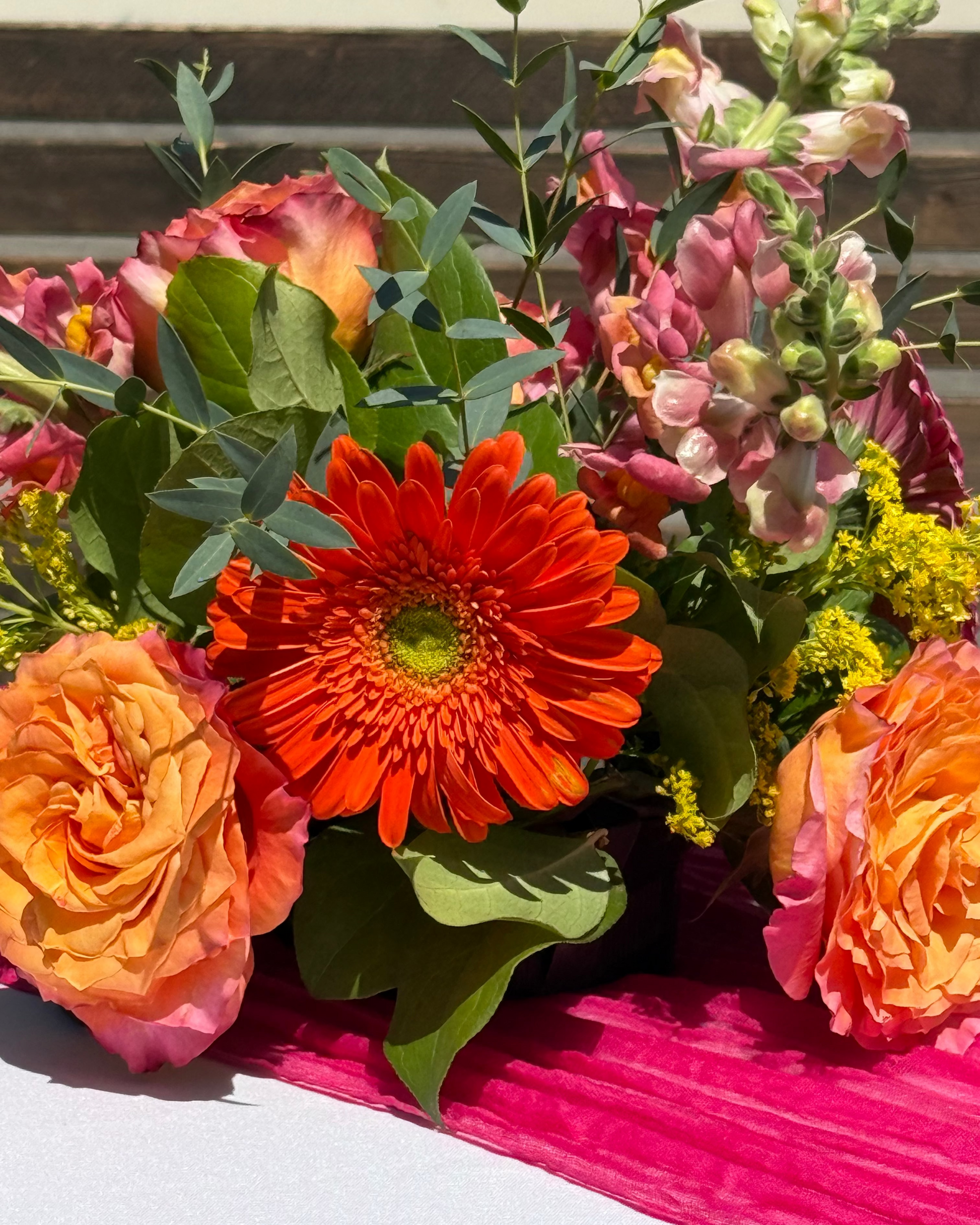 A vibrant floral arrangement featuring a bright orange gerbera daisy, peach roses, pink snapdragons, and green foliage, styled on a white surface with a bright pink pleated runner.