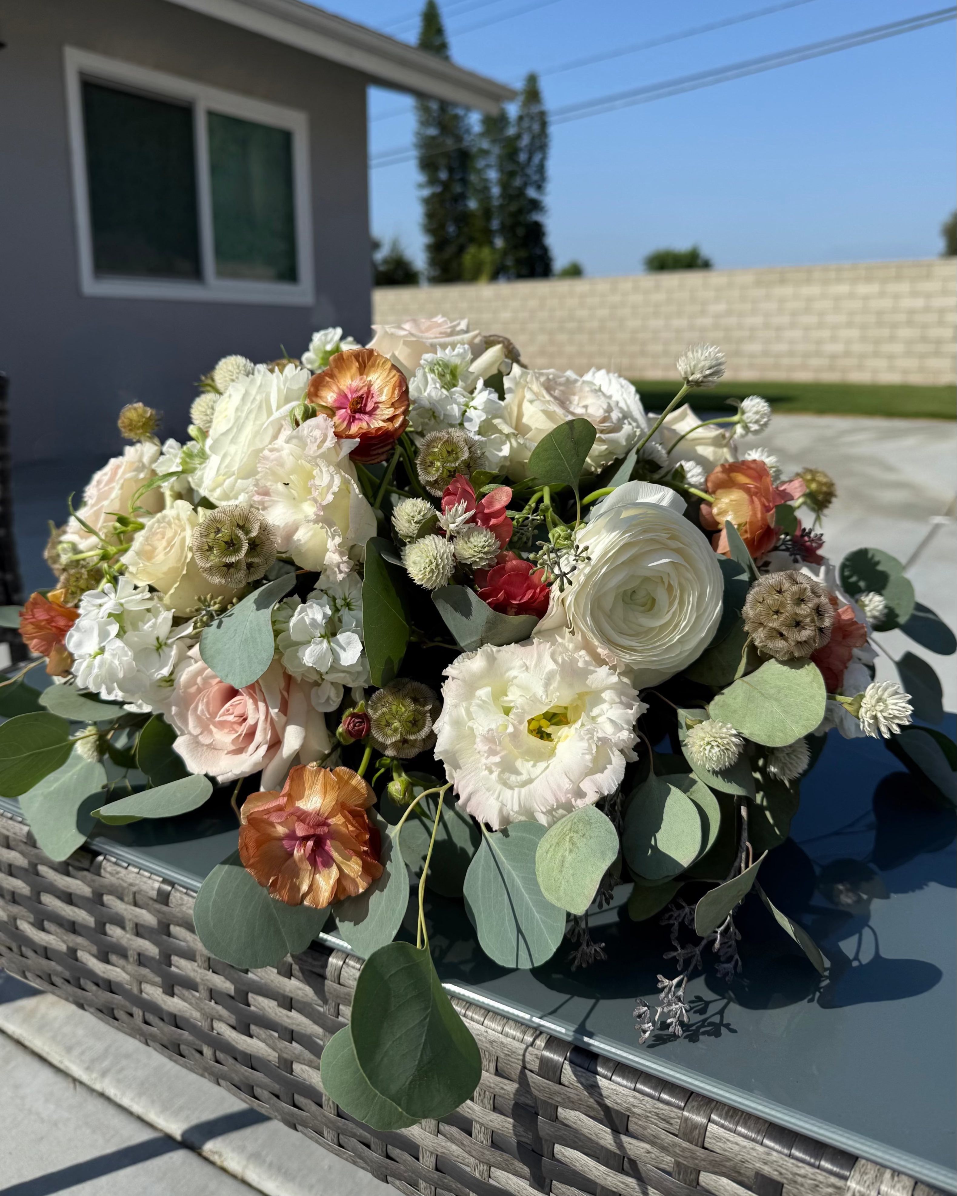 Bouquet of flowers in a woven basket on a concrete surface with a house and trees in the background.