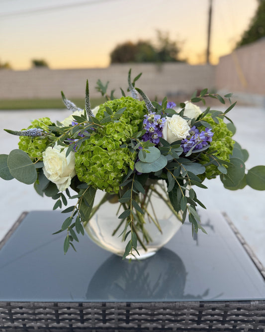 Superbloom Floral, Southern California Mobile Flower Shop bouquet of green and white flowers in a clear vase on a reflective surface with a blurred outdoor background.
