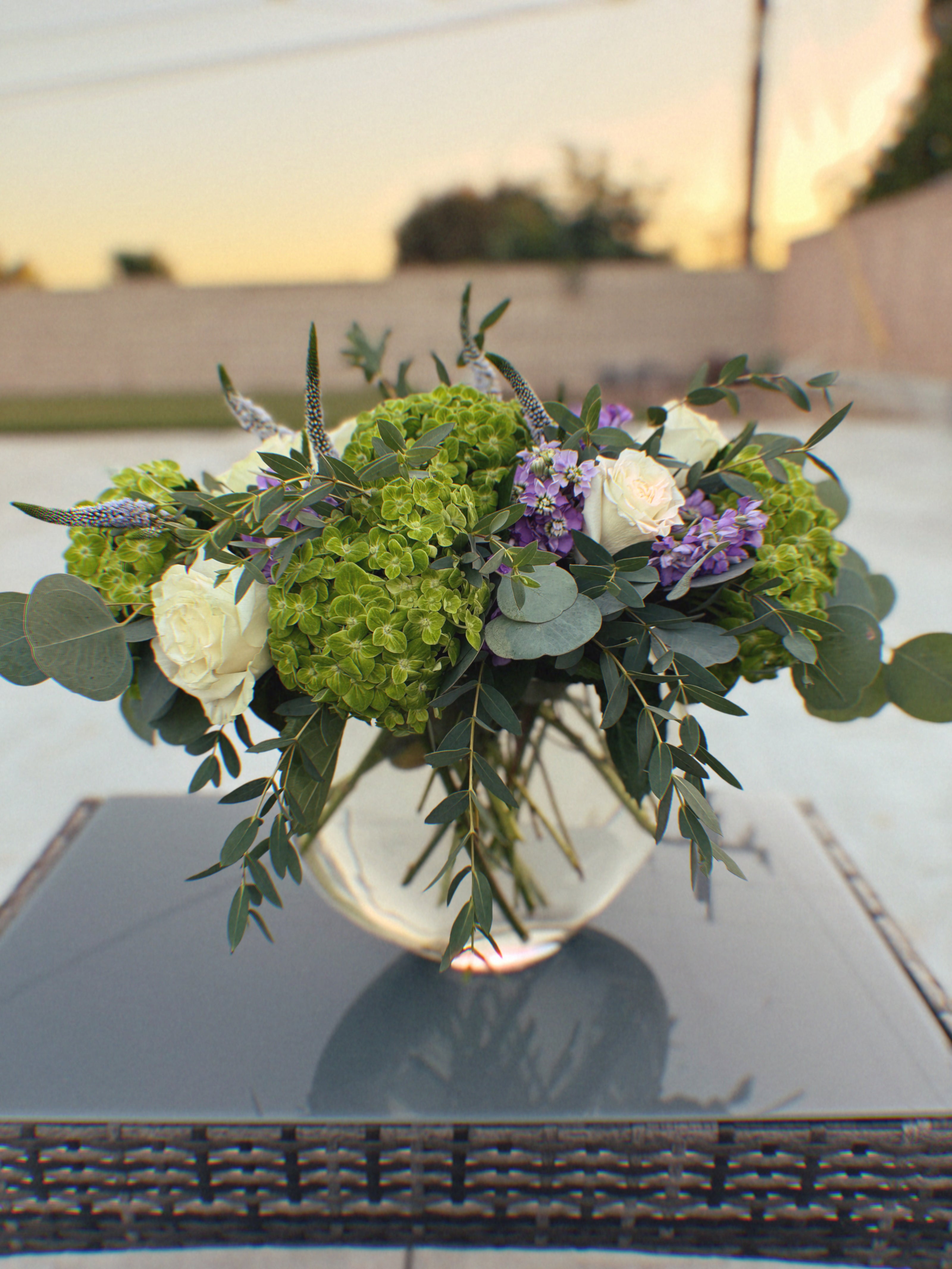 Floral arrangement with green hydrangeas, white roses, and purple flowers in a clear vase on a reflective surface.