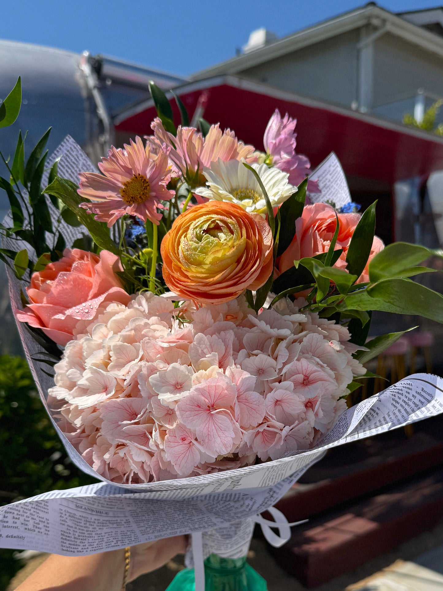 A vibrant flower bouquet outdoors. The bouquet features light pink hydrangeas, pink garden roses, pink cosmos and white gerbera daisies with fresh green leaves, wrapped in white newsprint paper.