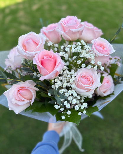 A bouquet of light pink roses and white baby's breath flowers with green foliage, held by a person wearing a blue sleeve against a blurred green grass background.