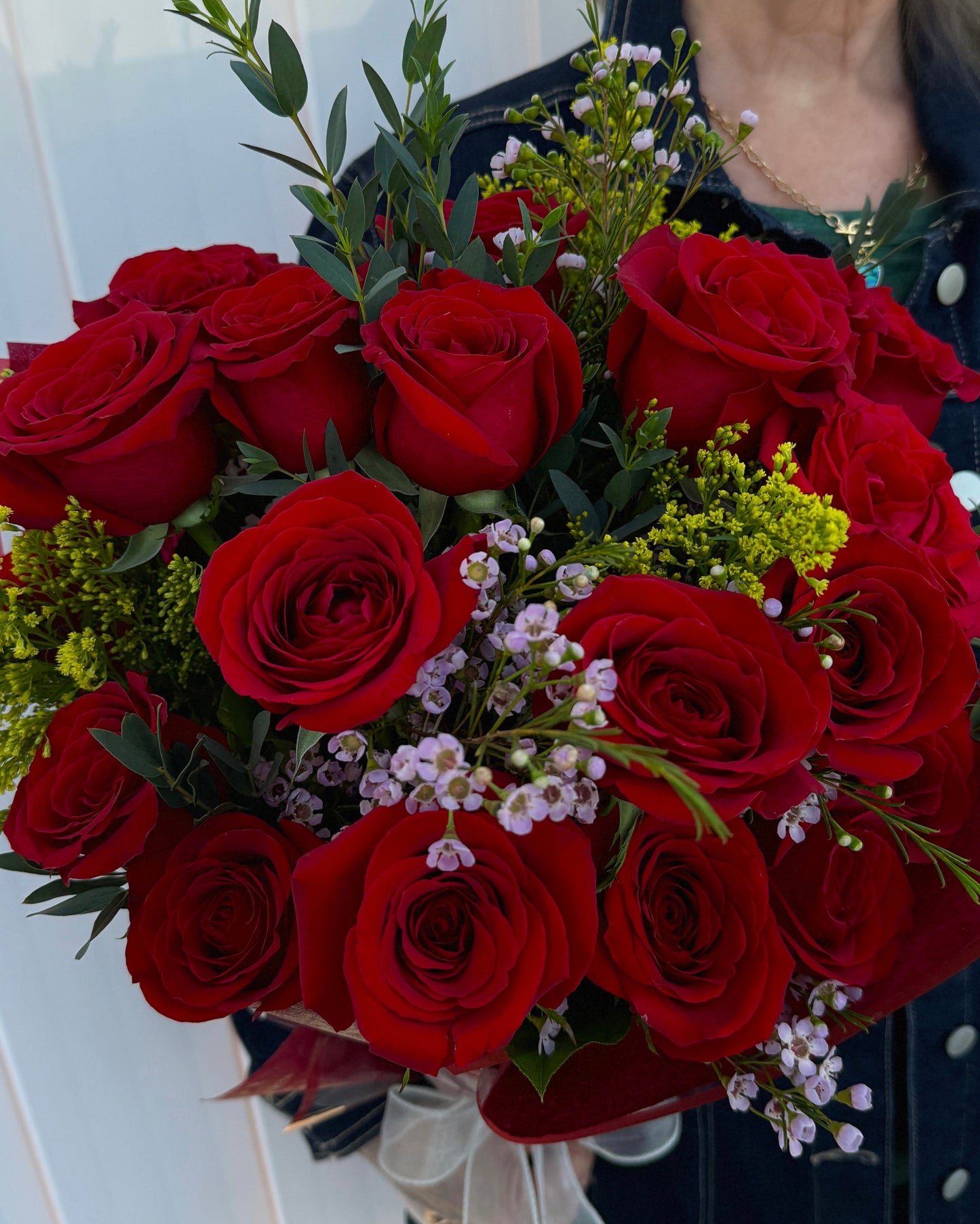 A close-up of a bouquet of red roses with green foliage and small white and yellow filler flowers, held by a person wearing a dark jacket.