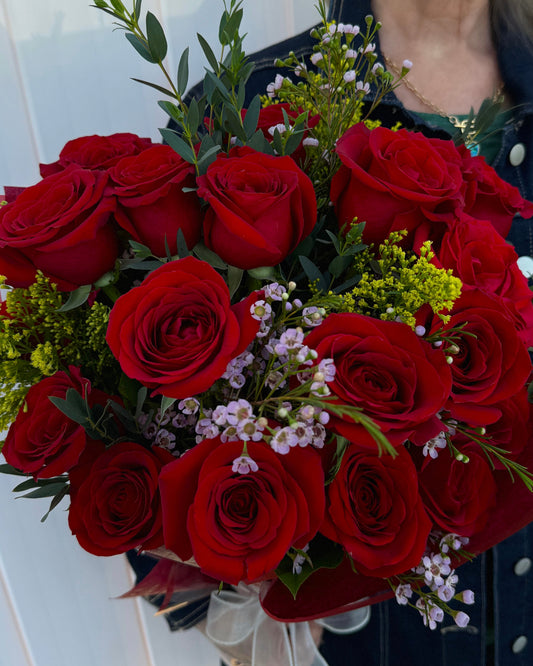 A close-up of a bouquet of red roses with green foliage and small white and yellow filler flowers, held by a person wearing a dark jacket.