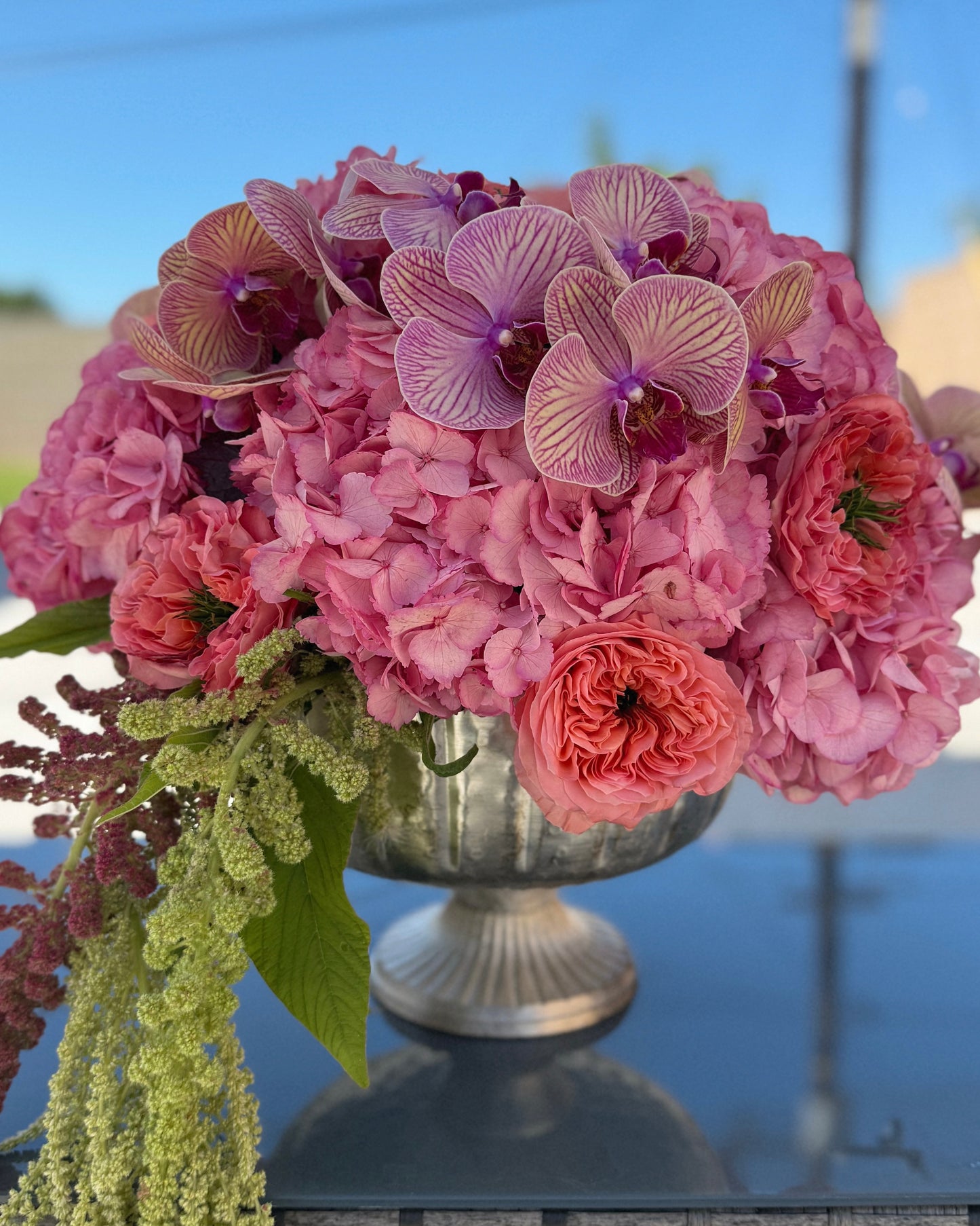 Bouquet of pink flowers in a silver vase on a reflective surface with a blurred outdoor background.