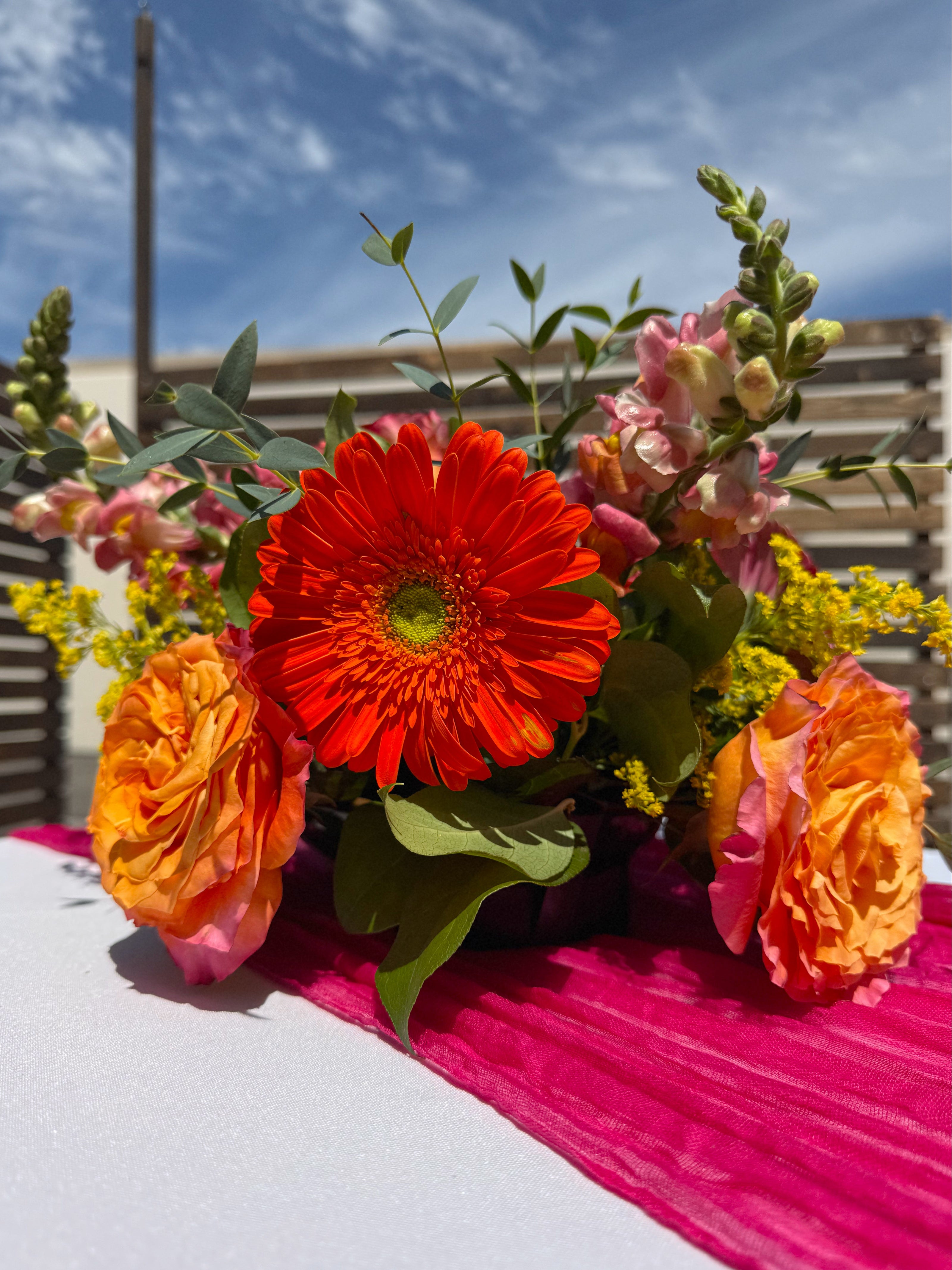 A vibrant floral centerpiece with a bright red-orange gerbera daisy, orange roses, pink snapdragons, and green foliage, arranged on a white table runner with a bright pink sash outdoors under a blue sky.