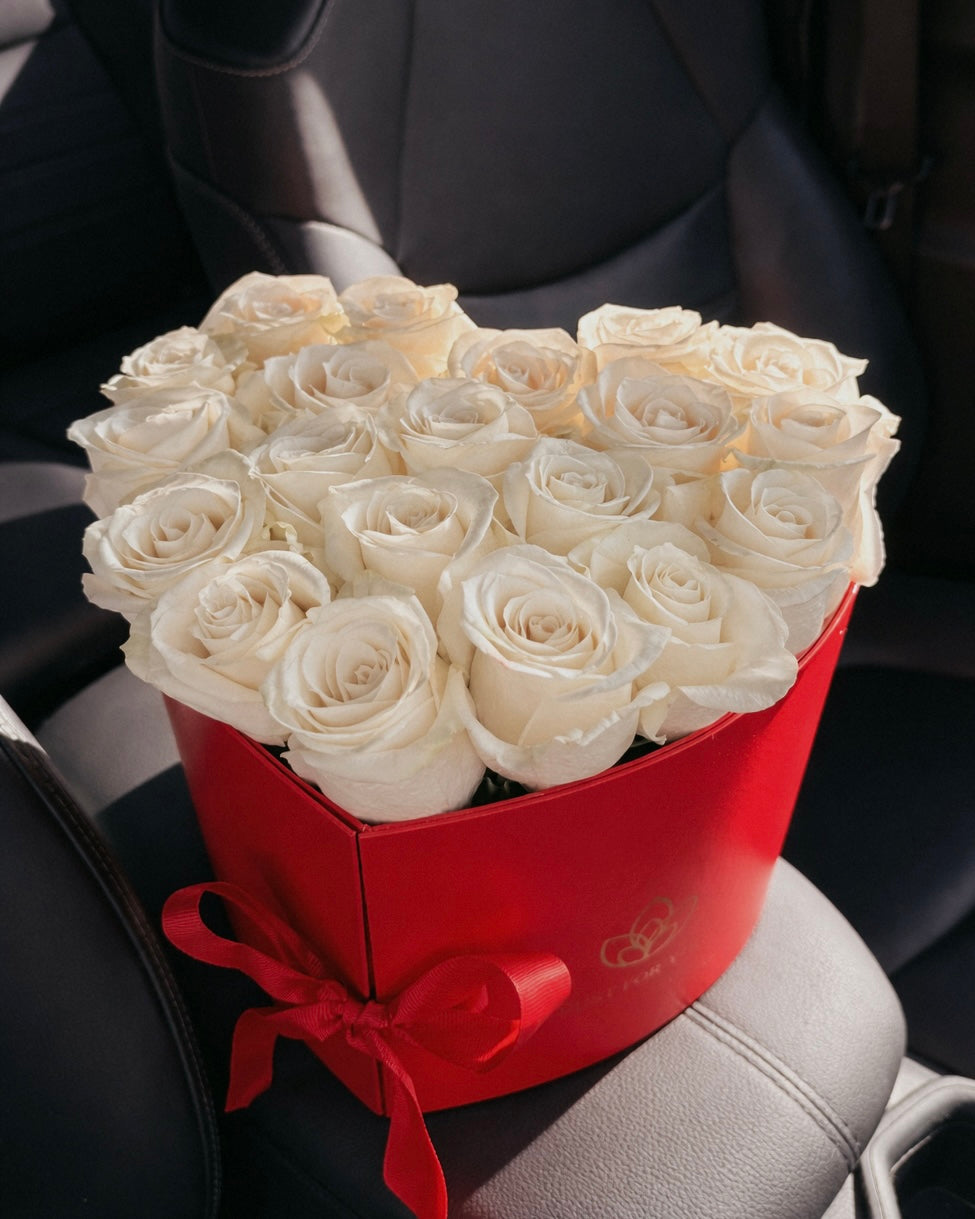 Heart-shaped box of white roses with a red bow on a car seat.