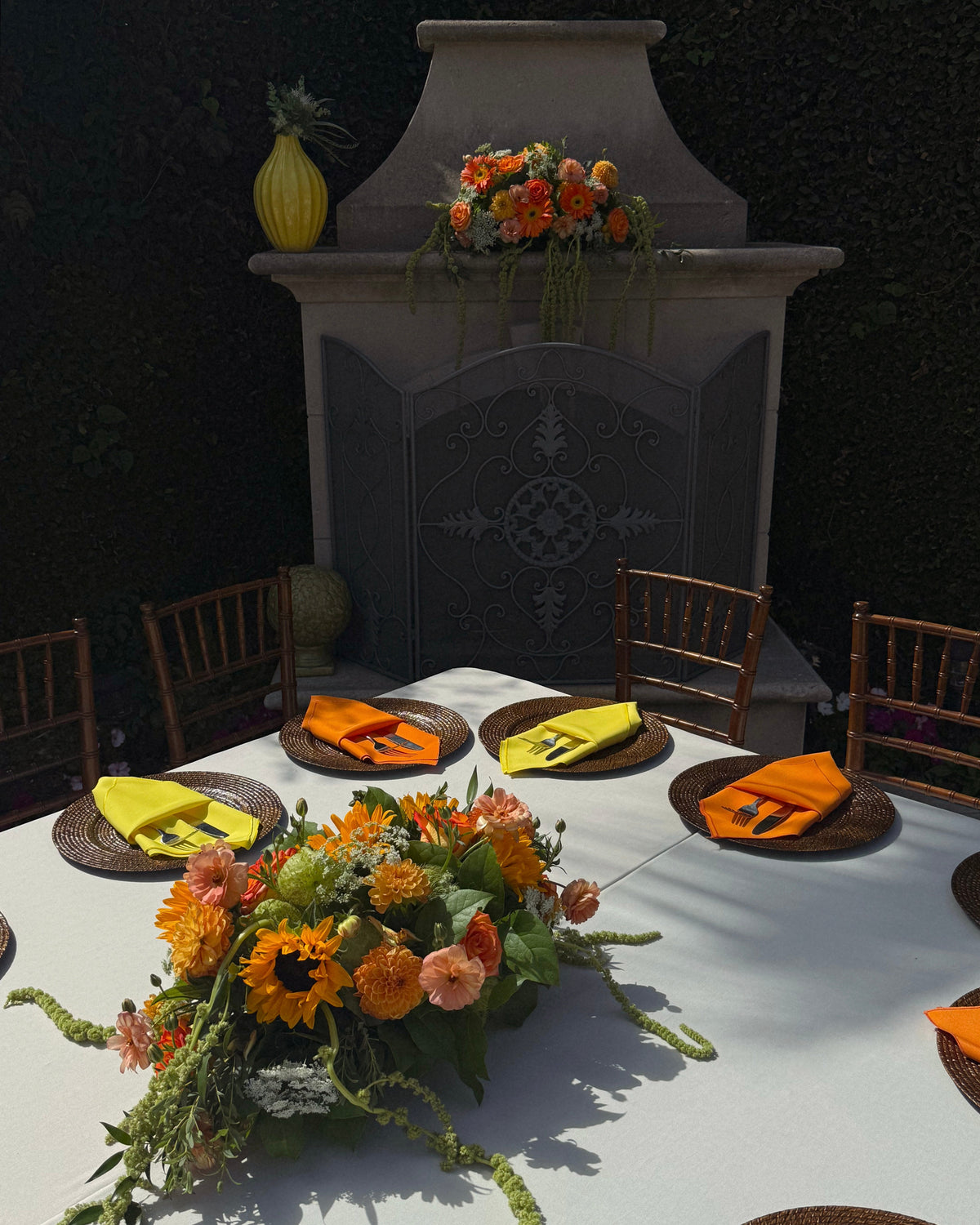 An outdoor dining table set with a large floral centerpiece, orange and yellow napkins, woven placemats, and wooden chairs in front of a stone fireplace feature.