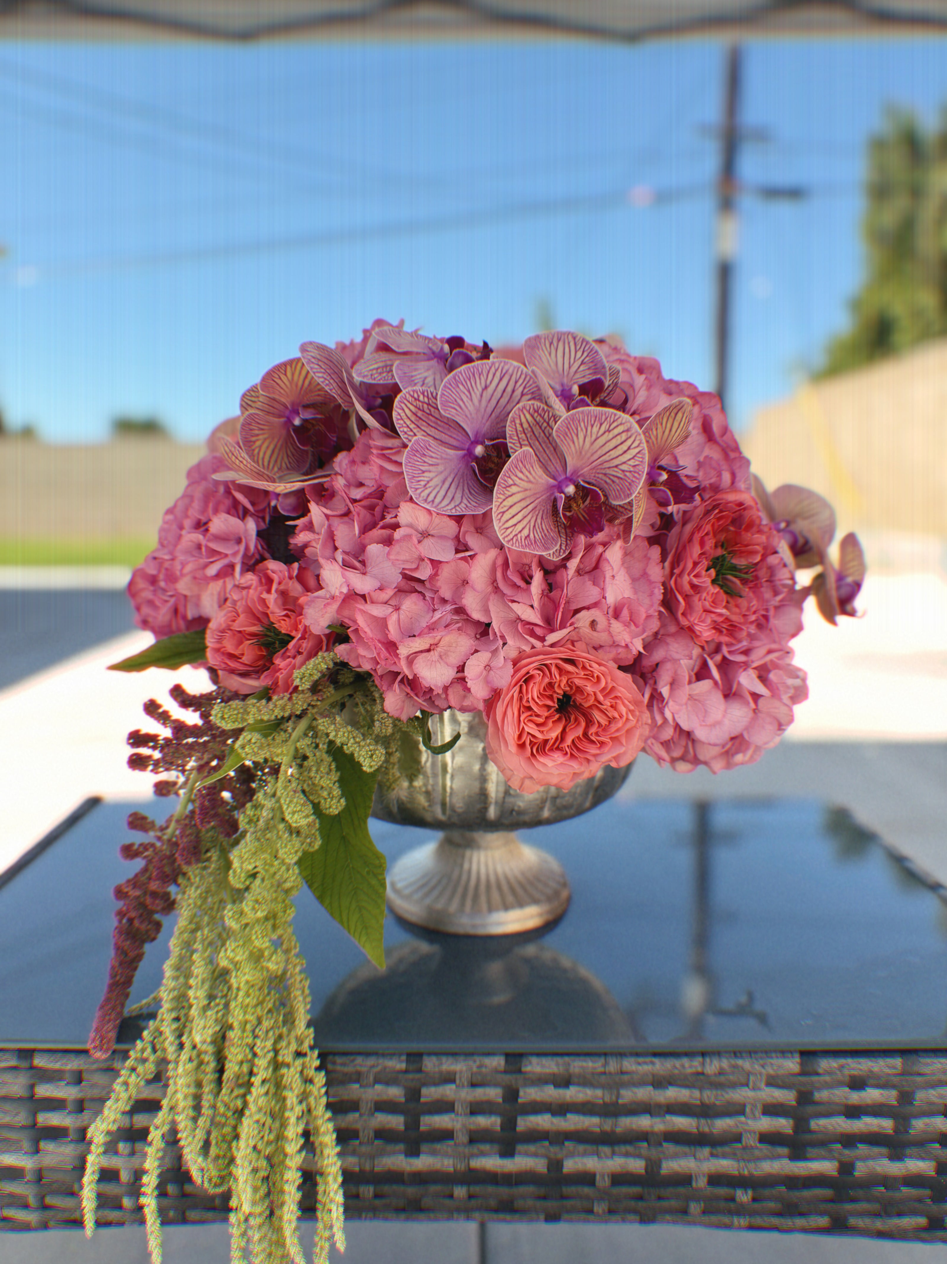 Bouquet of pink flowers in a silver vase on a reflective surface with a blurred outdoor background.
